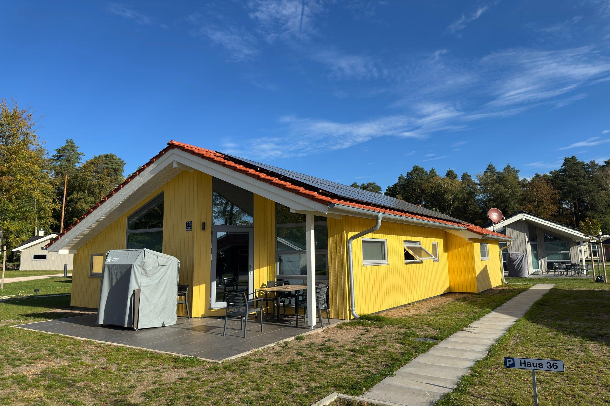 En gul bygning med rødt tag, et overdækket udendørs siddeområde og et skilt med teksten 'Haus 36'.