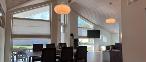 A spacious dining area with a high ceiling and large windows, a television mounted on the wall, and a cozy seating area with a fireplace.