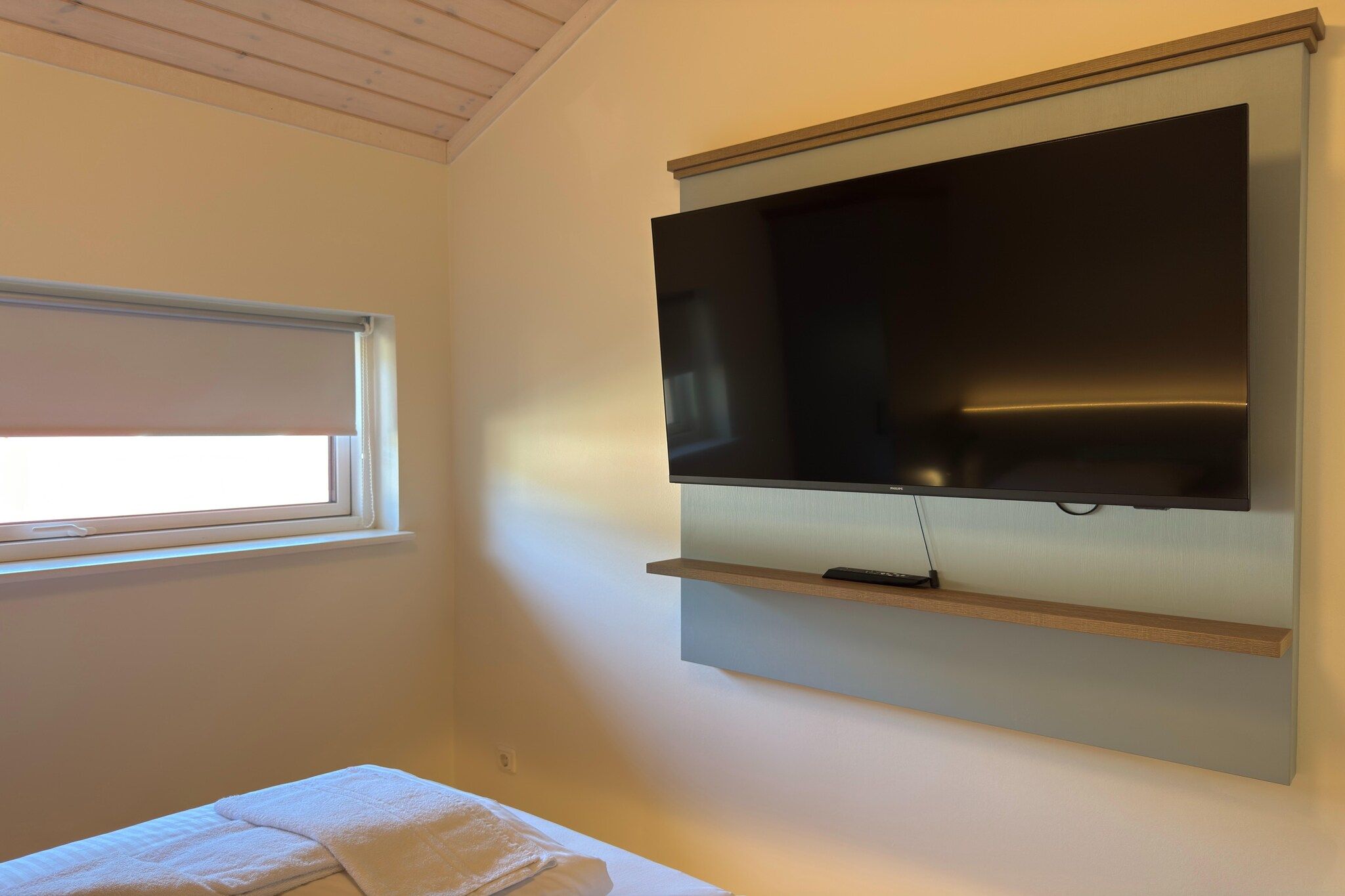 Et værelse med et fladskærms-tv på væggen, et vindue med en hvid markise, og et hvidt håndklæde på en bænk.