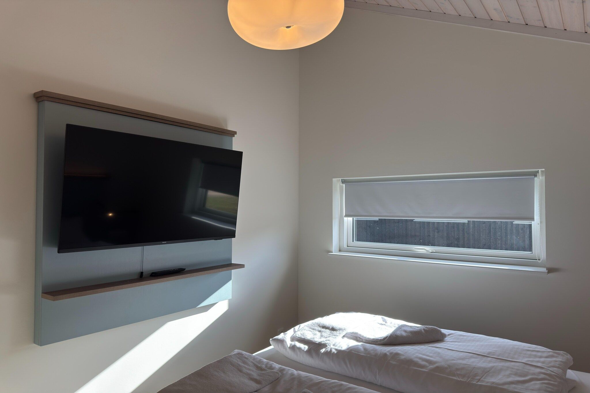 Une chambre avec un téléviseur fixé au mur, une fenêtre avec des stores et un lit avec des oreillers.