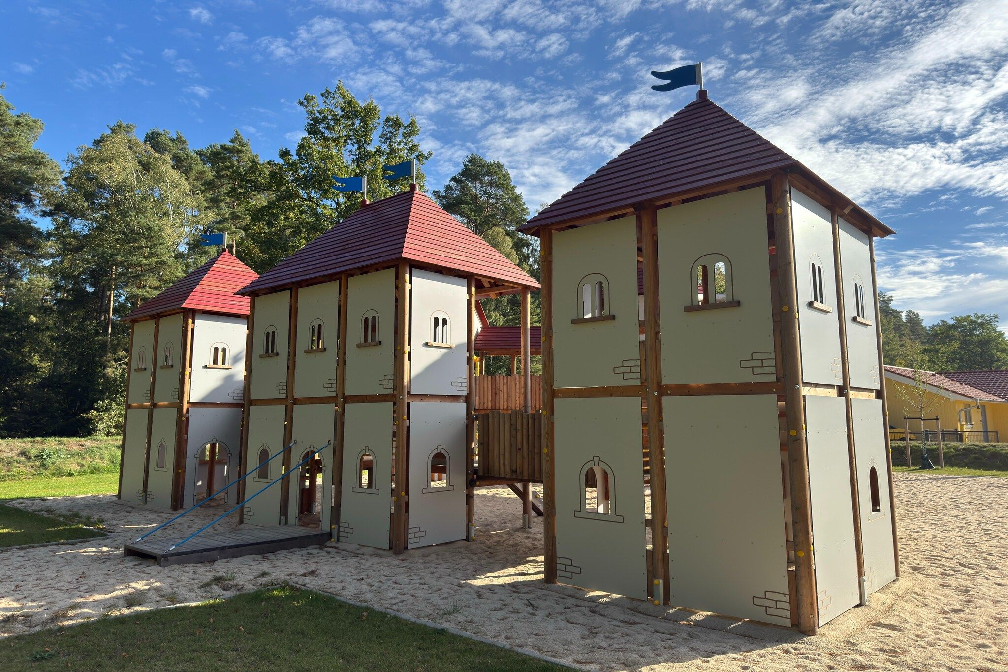 A playground with multiple play structures, including a slide and climbing areas, set on a sandy surface with trees in the background.
