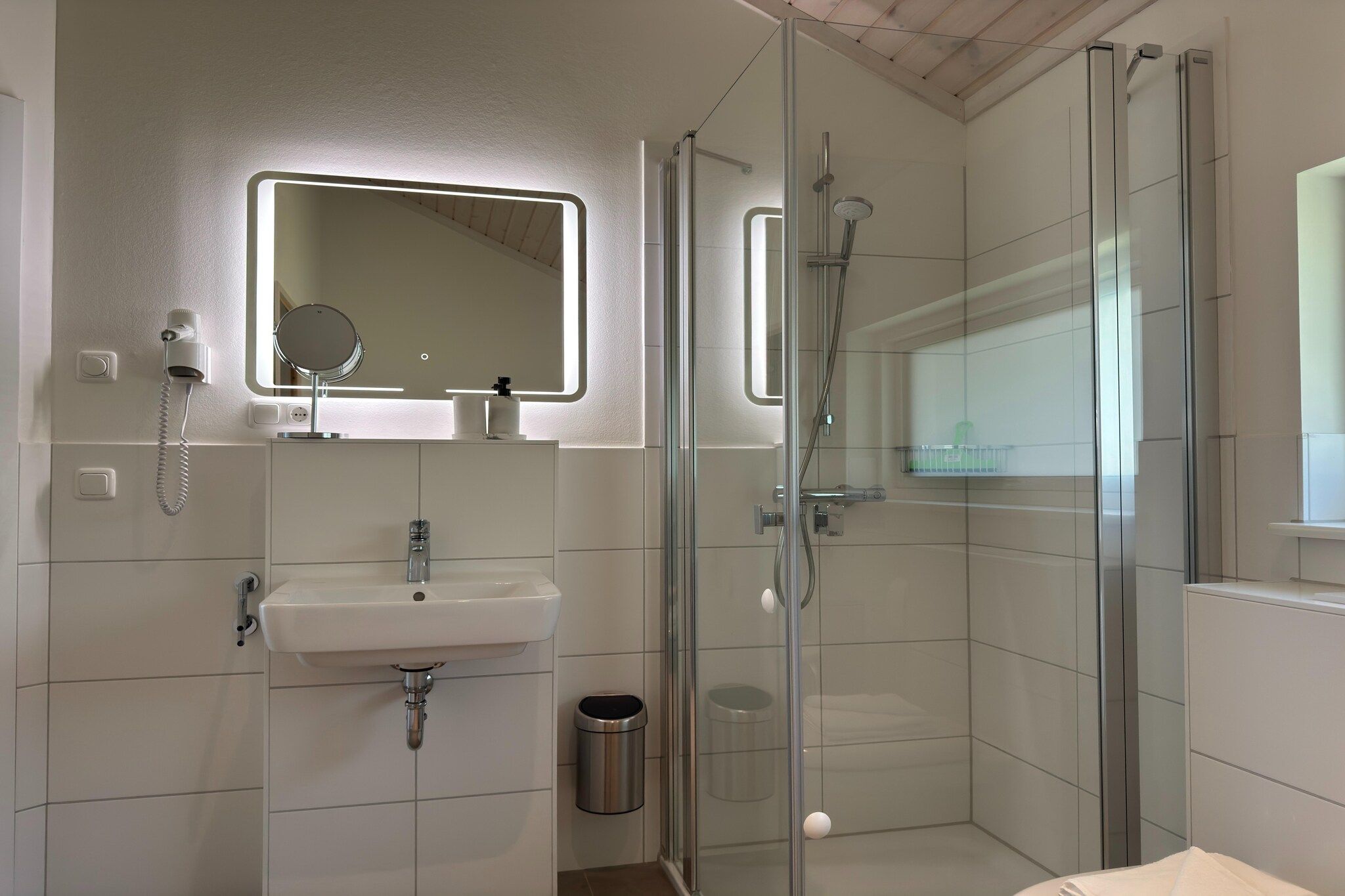 Ein modernes Badezimmer mit Duschkabine aus Glas, Waschbecken mit Spiegel darüber und einem Mülleimer.
