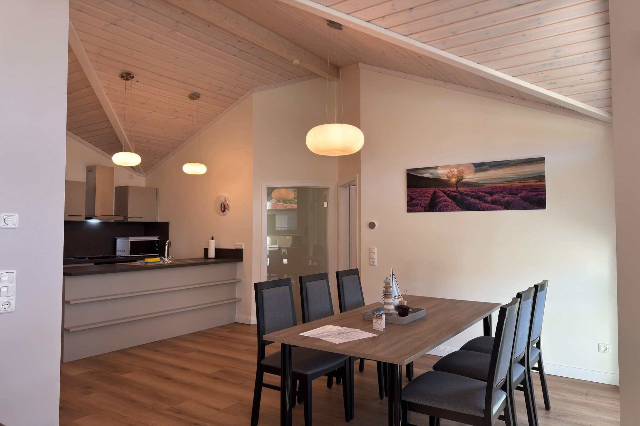Een eetgedeelte met een houten tafel en stoelen, een keuken op de achtergrond en een schilderij aan de muur.
