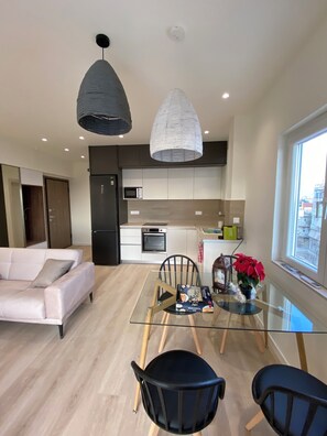 Ein modernes Wohnzimmer mit einem Glastisch, schwarzen Stühlen, einem weißen Sofa und einer Küche im Hintergrund.