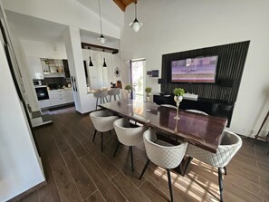 A modern kitchen with white cabinets, a dining area with a dark wooden table and white chairs, and a TV mounted on a black panel. - Beautiful Villa Near Etna and see (Trecastagni)
