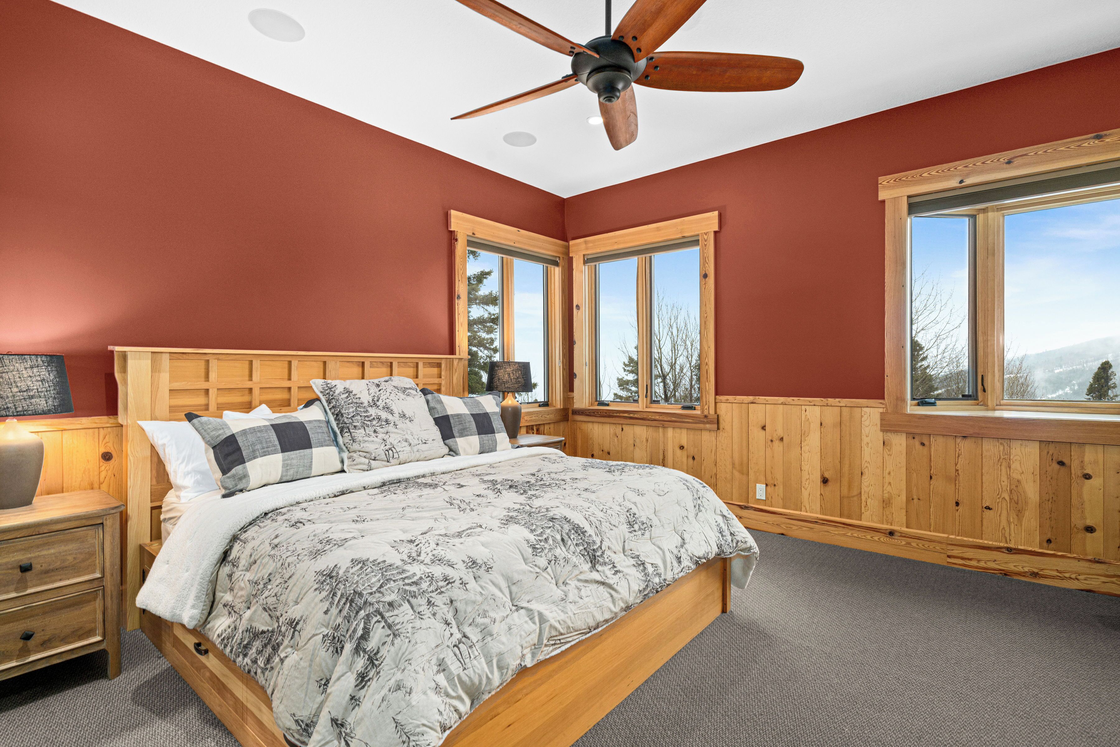 A bedroom with a wooden headboard, a bed with patterned bedding, a nightstand, a ceiling fan, and large windows with a view.