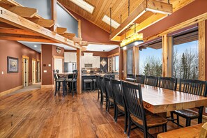 A spacious dining area with a large wooden table and chairs, a kitchen in the background, and a view of trees through large windows. - Panoramic Views of Lake Superior! Eagle Mountain Lodge at Lutsen (Lutsen)