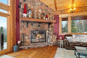 A stone fireplace with a log fire, a leather sofa, and a large window overlooking a forest. - Panoramic Views of Lake Superior! Eagle Mountain Lodge at Lutsen (Lutsen)