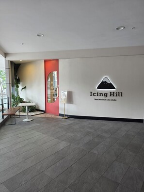 Um interior moderno com o logotipo de 'Icing Hill Your Personal cake studio' na parede, uma porta vermelha e uma área de estar com uma mesa e um banco.