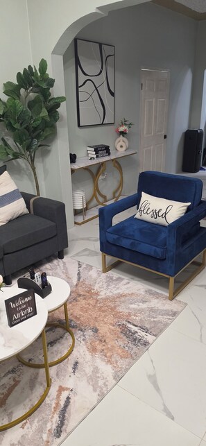 Ein modernes Wohnzimmer mit einem blauen Sessel, einem dunklen Sofa, einem runden Couchtisch und einem Schild mit der Aufschrift 'Willkommen bei Airbnb'.