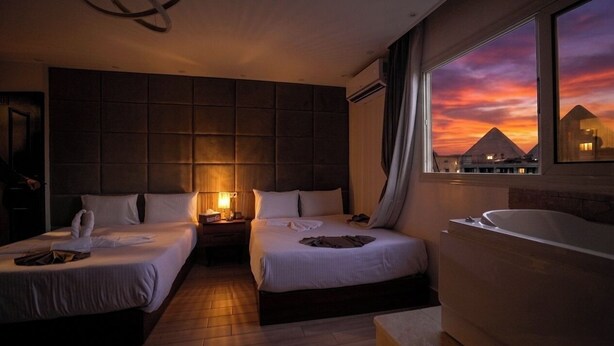 Habitación de hotel con dos camas, un ventanal con vistas al atardecer, una bañera y una lámpara moderna.