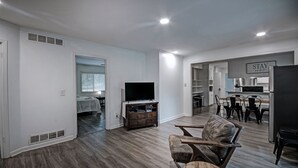 A modern living room with a flat-screen TV, wooden flooring, a dining area with a table and chairs, and a kitchen with a refrigerator and cabinets. - 7BR Workforce Stay Near I-55 & I-220 (Jackson)