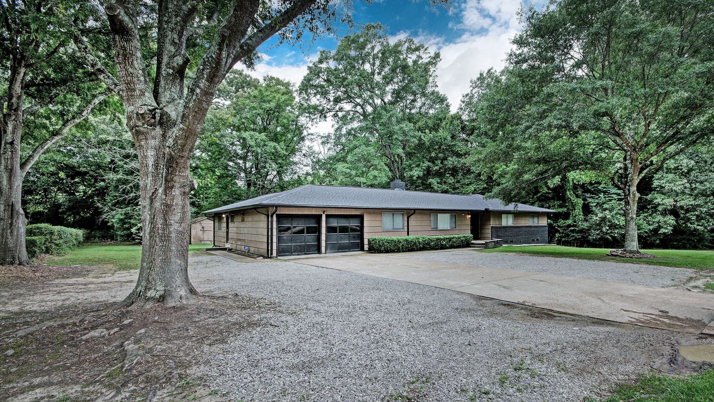 A single-story house with a garage, surrounded by trees and a gravel driveway.
