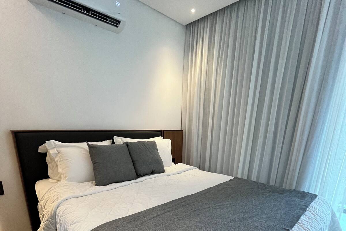 Un dormitori d'hotel modern amb un llit gran, un capçal, una finestra amb cortines i una unitat de climatització a la paret.