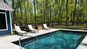 Una piscina rectangular con una escalera metálica, rodeada por un solárium de concreto. Hay sillones de descanso y una pequeña estructura de madera con una ventana.