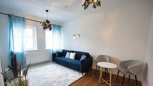 Un salon avec un canapé bleu, deux fauteuils, un tapis blanc, un plancher en bois, une fenêtre avec des rideaux bleus et un luminaire au plafond.