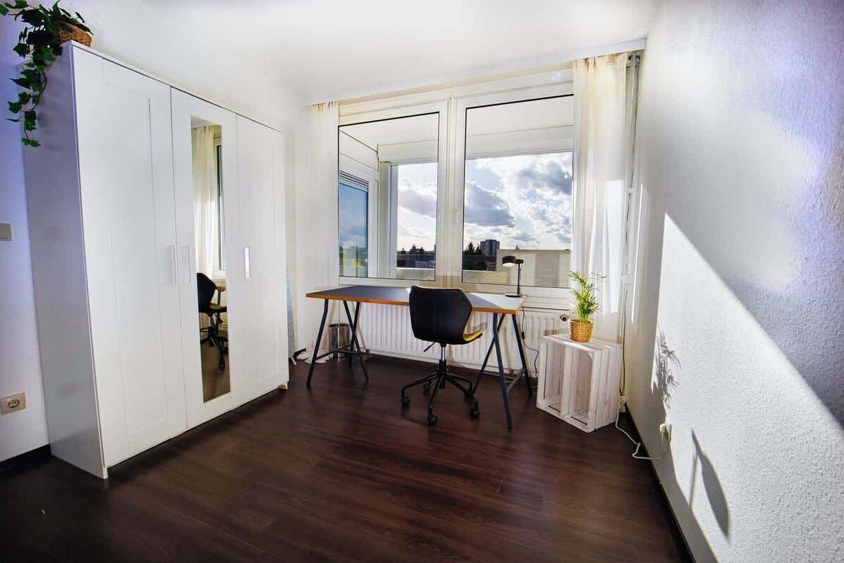 A room with a wooden floor, a desk with a chair, a radiator, and a window with a view of the sky.