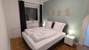 Een net opgemaakt bed met witte lakens, een nachtkastje met een lamp en een raam met gordijnen.
