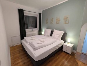 Ein ordentlich bezogenes Bett mit weißen Leinen, ein Nachttisch mit Lampe und ein Fenster mit Vorhängen.