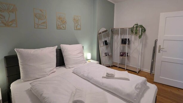 Een net opgemaakt bed met witte lakens, een nachtkastje met een lamp en een kunstwerk aan de muur.