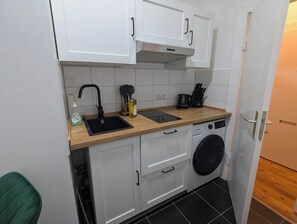 Eine kleine Küche mit weißen Schränken, einer Holzarbeitsplatte, einem Spülbecken, einer Waschmaschine und einer Kaffeemaschine.