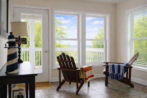 A room with a view of trees and a lake, a wooden chair, a small table with a lamp, and a bench with a blanket. - Aqua Breeze/Lakefront, Hot Tub, Fire Pit, 3 Decks (Michigan City)