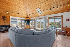 Ein modernes Wohnzimmer mit einem großen grauen Ecksofa, Holzbalkendecke, großen Fenstern und einem Kamin.