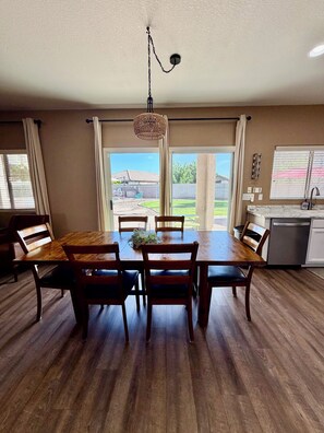 Un espace repas avec une table et des chaises en bois, un luminaire suspendu et une vue sur l’extérieur grâce à de grandes fenêtres.