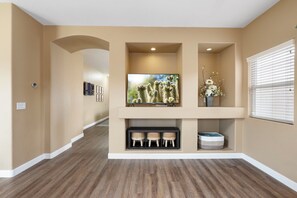 A hallway with a flat-screen TV mounted on a wall, a small shelf with decorative items, and a window with blinds. - Desert Family Oasis, Pool, Spa & Kids’ Paradise (Avondale)