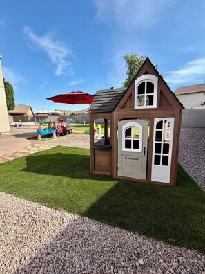Une petite maisonnette en bois, dotée d’une porte et de fenêtres, entourée d’une pelouse et agrémentée d’un parasol rouge.
