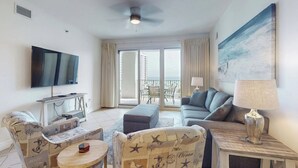 A living room with a flat-screen TV, a sofa, a coffee table, and a balcony with a view of a building. - 🏖️Gulf + Pool Views | Walk to Beach | 2BR Ariel Dunes I 609 🏖️ (Miramar Beach)