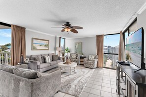 A modern living room with a ceiling fan, a large window, and a flat-screen TV. - Walk to Beach🌴 2 Pools + Hot Tub☀️Mainsail 334 (Miramar Beach)