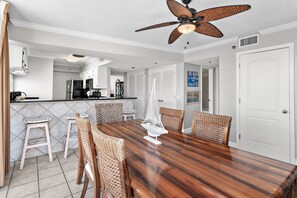 A dining area with a wooden table, wicker chairs, a ceiling fan, and a kitchen in the background. - Walk to Beach🌴 2 Pools + Hot Tub☀️Mainsail 334 (Miramar Beach)