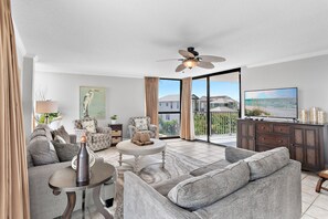 A spacious living room with a large sofa, a coffee table, and a television. There is a ceiling fan, a painting on the wall, and a view of the outside through large windows. - Walk to Beach🌴 2 Pools + Hot Tub☀️Mainsail 334 (Miramar Beach)