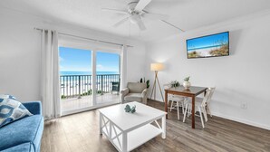 Ein Wohnzimmer mit Balkon, Deckenventilator, Esstisch mit Stühlen und einem an der Wand befestigten Fernseher.