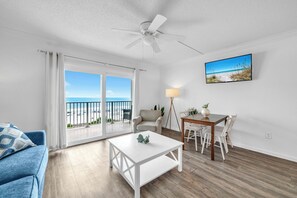 Ein Wohnzimmer mit Balkon, Deckenventilator, Esstisch mit Stühlen und einem an der Wand befestigten Fernseher.