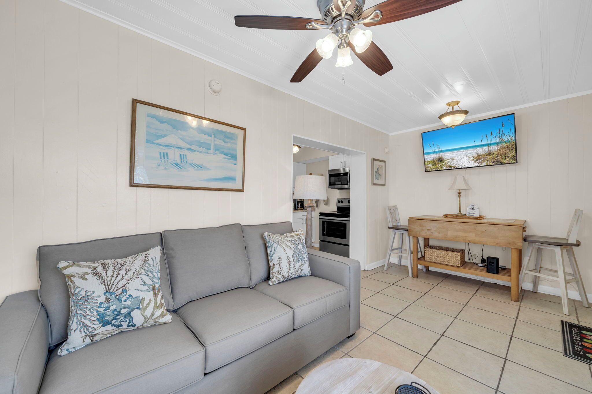 A living room with a grey sofa, a ceiling fan, a wooden table, and a kitchen in the background.