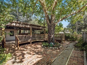 Eine Holzdielen-Terrasse mit einer Pergola, einem Pflasterweg und einem großen Baum.