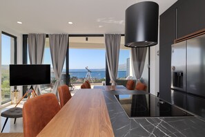 Ein moderner Essbereich mit einem langen Tisch, Stühlen, einem Flachbildfernseher und Blick auf das Meer.