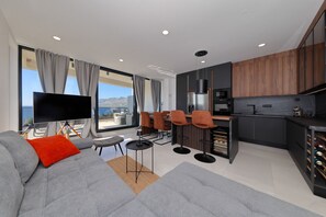 Ein stilvoller Wohnbereich mit Sofa, Fernseher, Esstisch und Stühlen, eine Küche mit dunklen Schränken und Meerblick.