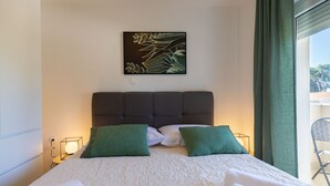 Hotellihuone, jossa on sänky, kaksi vihreää tyynyä, sängynpääty, seinälle ripustettu taideteos ja ikkuna, jossa on verhot.