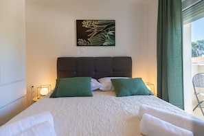 Ein Hotelzimmer mit einem Bett, zwei grünen Kissen, einem Kopfteil, einem Wandbild und einem Fenster mit Vorhängen.