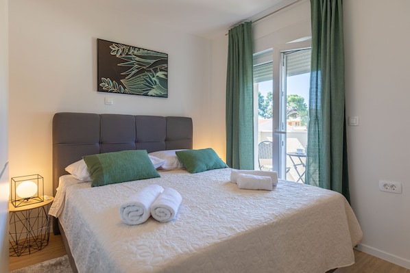Ein ordentlich bezogenes Bett mit grünen Kissen, ein an der Wand befestigtes Kunstwerk und ein Blick durch ein mit Vorhängen drapiertes Fenster.