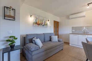 A modern living room with a grey sofa, a small table with a bonsai, a painting of sailboats, a wooden door, and a kitchen area with a sink and cabinets. - Apartment Klanac 2 with Two Balconies (Zadar)