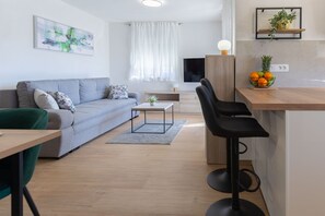 A modern living room with a grey sofa, a small coffee table, a green chair, a black bar stool, and a kitchen area with a fruit bowl. - Apartment Klanac 1 with Balcony (Zadar)