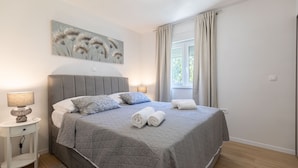 Un lit bien fait, recouvert d’une couverture grise, avec deux serviettes enroulées et une lampe de chevet posée sur une table de chevet blanche.