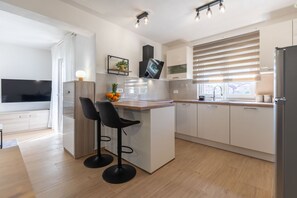 A modern kitchen with a central island, bar stools, a flat-screen TV, and a large window with blinds. - Apartment Klanac 1 with Balcony (Zadar)