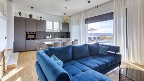 A modern living room with a blue sectional sofa, a dining area, a kitchen, and a large window with a view. - Villa Flokica with Heated Pool (Maslenica)
