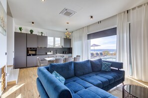 A modern living room with a blue sectional sofa, a dining area, a kitchen, and a large window with a view. - Villa Flokica with Heated Pool (Maslenica)
