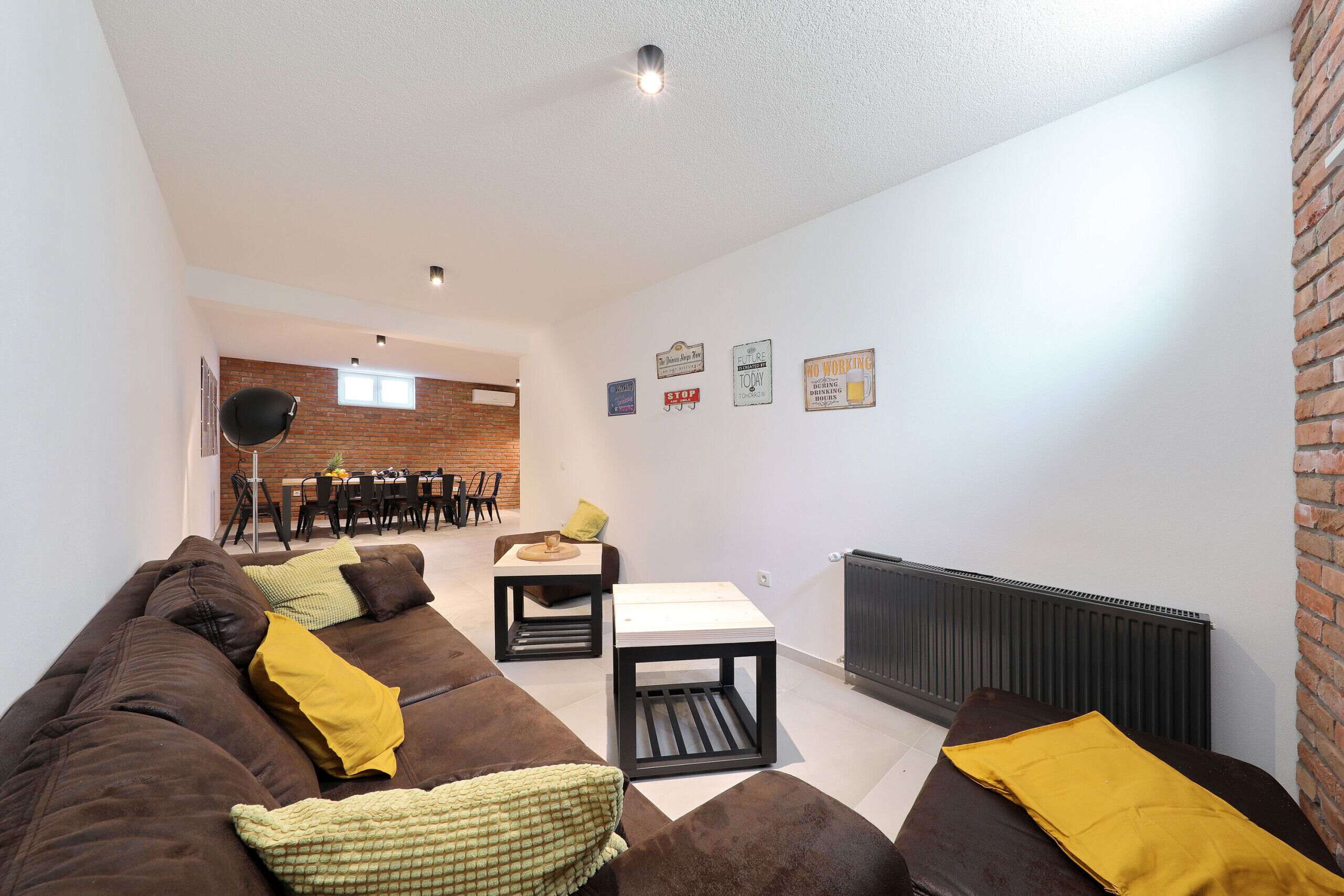 A living room with a brown sofa, a small table, and a radiator. There are chairs and a dining table in the background.